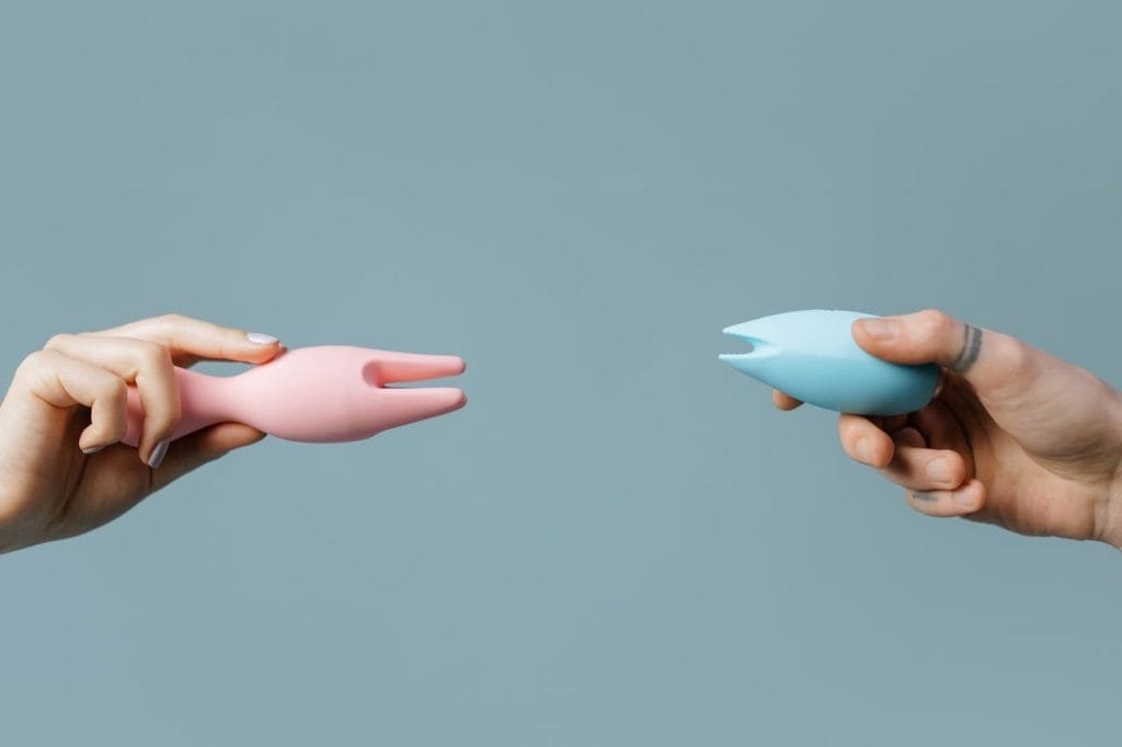 Two hands holding pink and blue silicone vibrators against a pastel background.